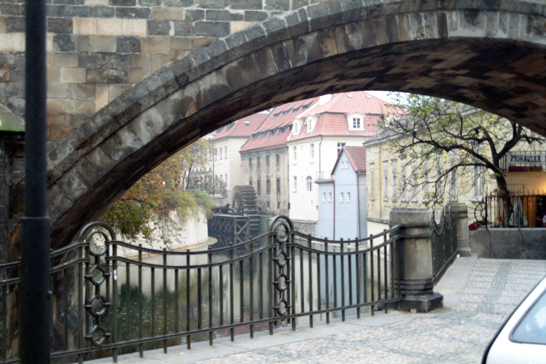 A stone arch bridge frames a quaint canal scene with historic buildings and a tree in Prague.