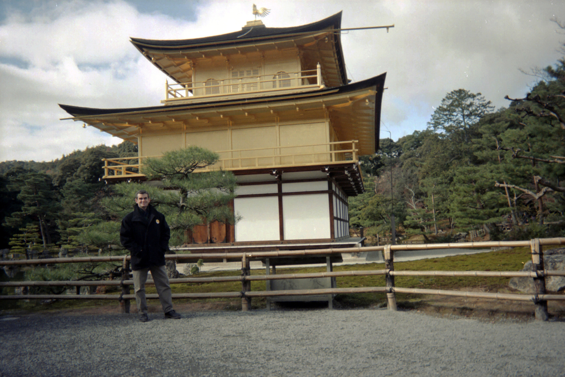 A person stands in front of the Kinkaku-ji, the Golden Pavilion, surrounded by lush greenery.